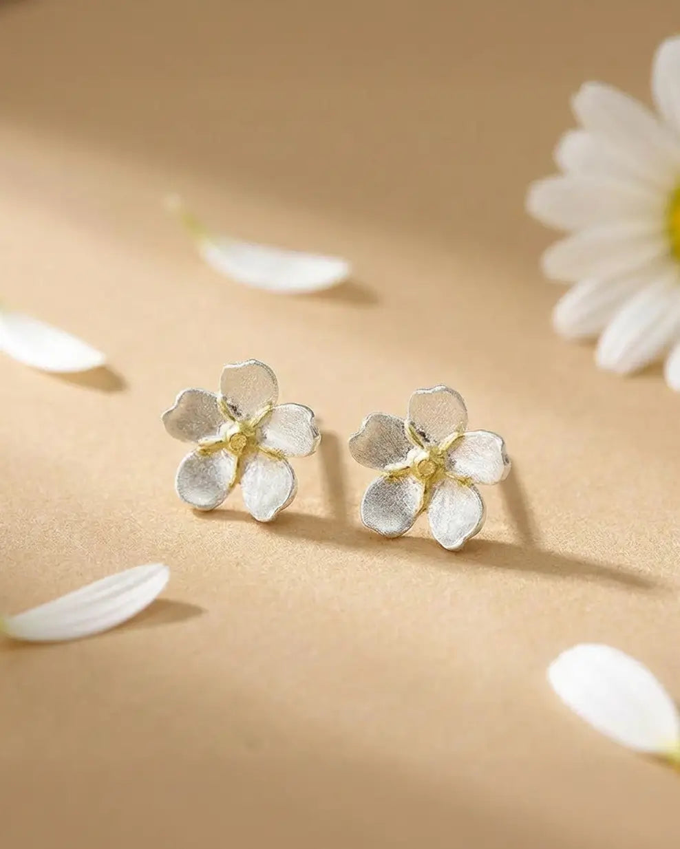 Boucles d’oreilles pâquerette en argent avec cœur doré et émail blanc, posées sur une surface claire avec des pétales, ambiance douce et lumineuse.