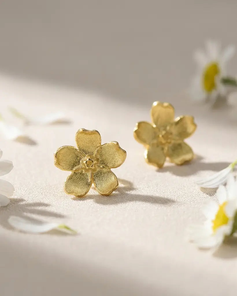 Boucles d’oreilles pâquerette en argent doré avec émail blanc, posées sur une surface claire avec des fleurs, ambiance douce et naturelle.