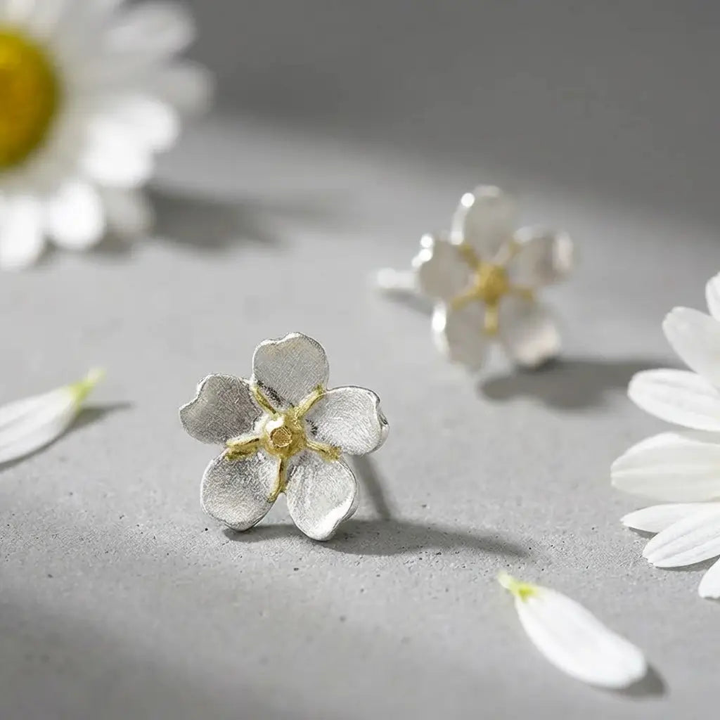 Boucles d’oreilles pâquerette en argent doré avec émail blanc et cœur doré, posées sur une surface claire avec des fleurs, ambiance douce et naturelle.