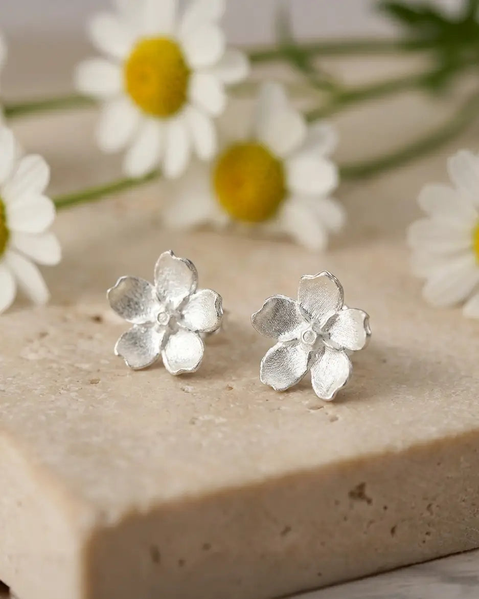 Boucles d’oreilles pâquerette en argent avec émail blanc, posées sur une surface naturelle avec des fleurs, ambiance douce et poétique.