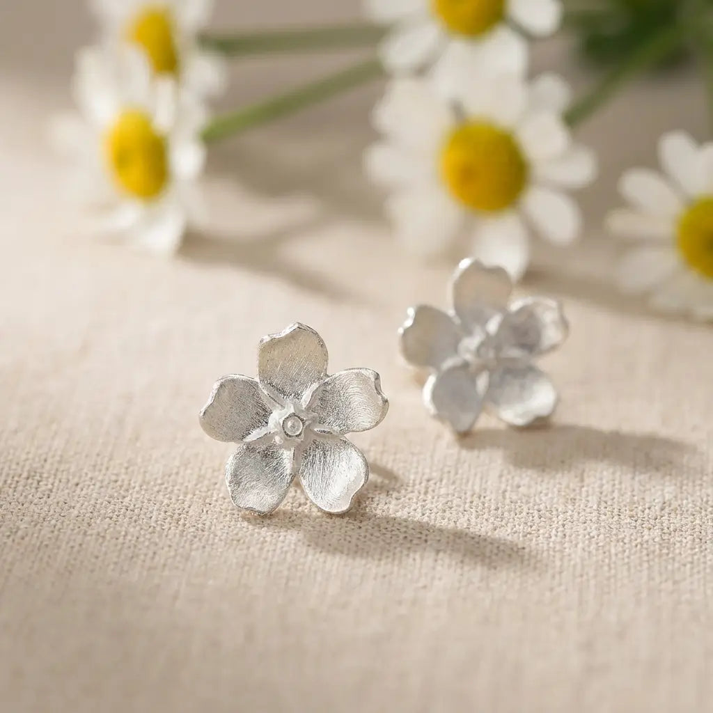 Boucles d’oreilles pâquerette en argent 925 avec émail blanc, posées sur une surface claire avec des fleurs, ambiance douce et naturelle.