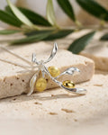 Collier branche d’olivier en argent et doré posé sur une surface minérale, bijou inspiré de la nature et de la paix.