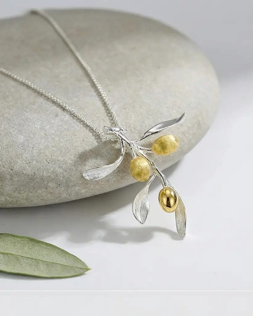 Collier branche d’olivier en argent et doré présenté à plat sur un galet, mettant en valeur les feuilles et olives dorées.