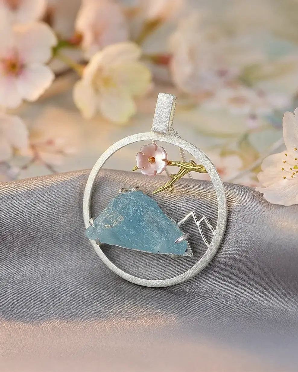 Médaillon floral du sommet en argent et or avec pierre bleue, posé dans une mise en scène poétique et florale.