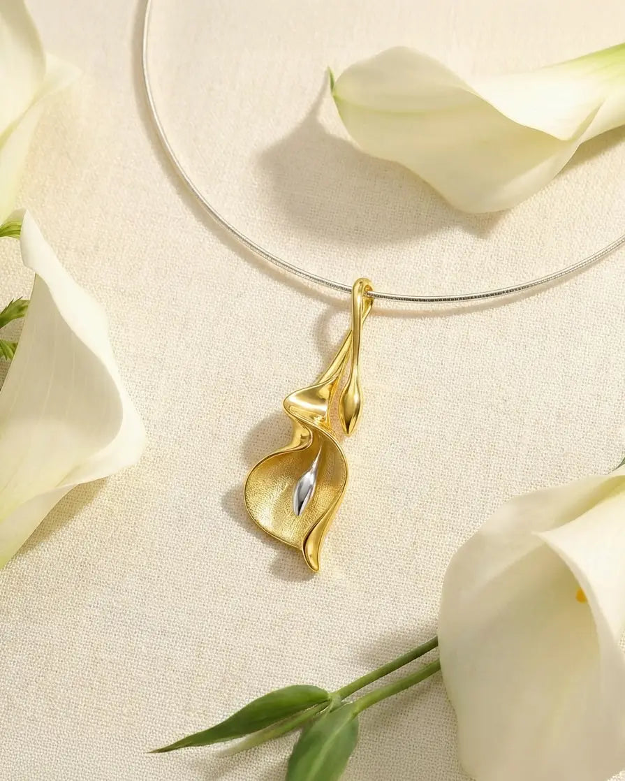 Pendentif arum en argent 925 avec finition dorée, posé sur un fond clair avec des fleurs blanches, ambiance douce et naturelle.